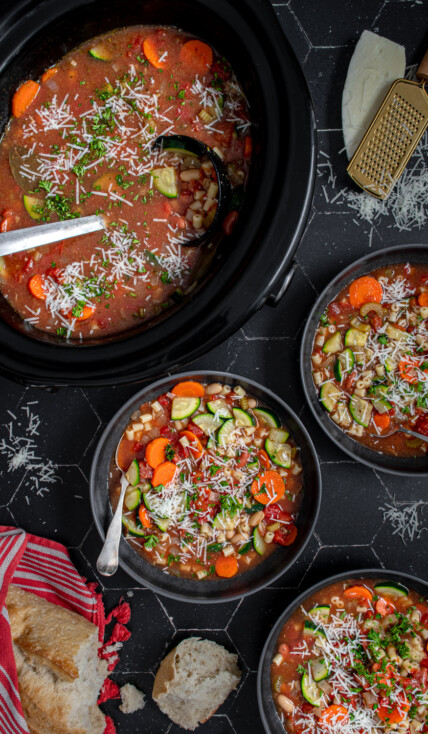 Table top with black bowls and crockpot filled with minestrone soup and garnished with parmesan cheese.
