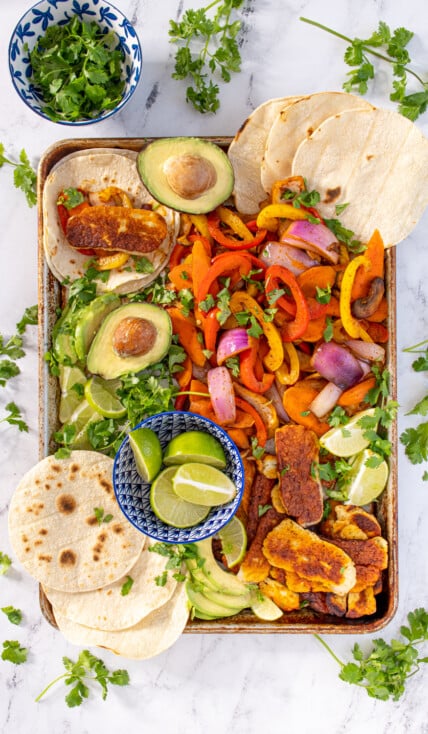 Overhead view of a tray filled with veggie halloumi fajita makings with tortillas, avocados, and lime wedges with everything garnished with cilantro.