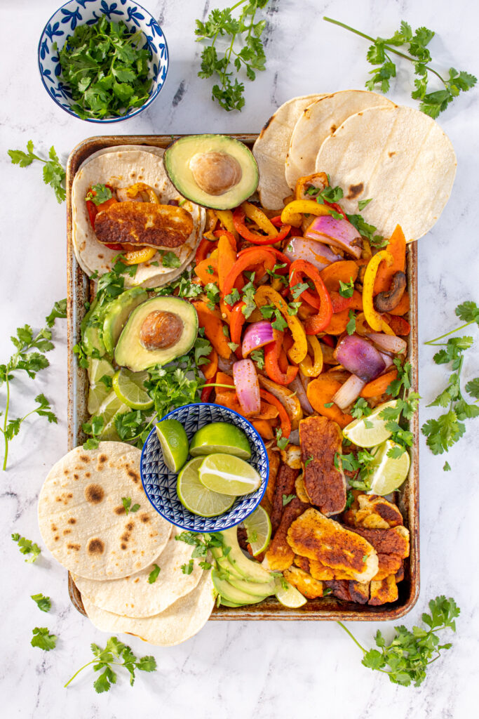 Overhead view of a tray filled with veggie halloumi fajita makings with tortillas, avocados, and lime wedges with everything garnished with cilantro.