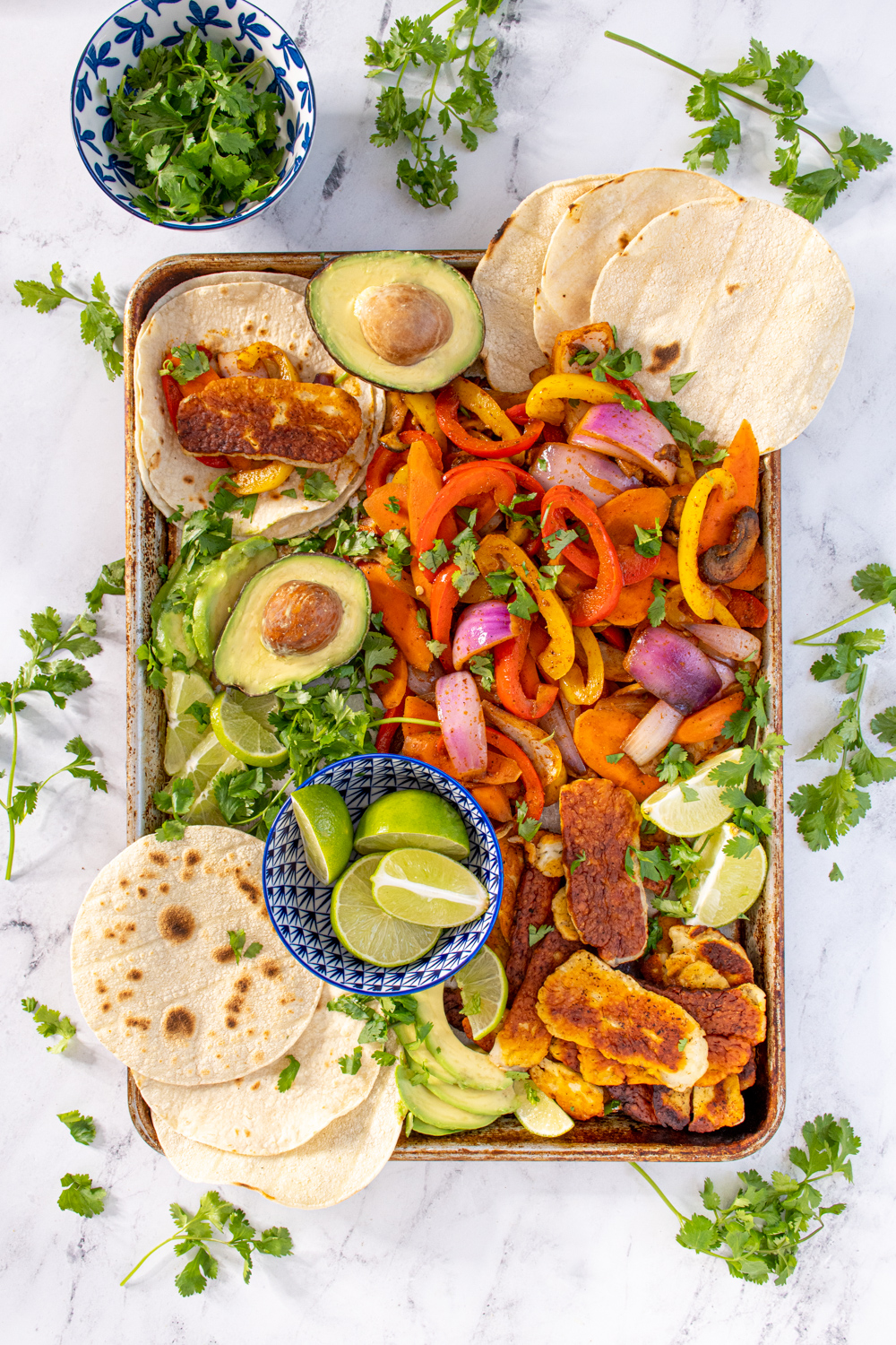 Overhead view of a tray filled with veggie halloumi fajita makings with tortillas, avocados, and lime wedges with everything garnished with cilantro.