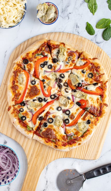 A homemade veggie pizza cut into pieces on a wooden pizza peel surrounded by pizza toppings.