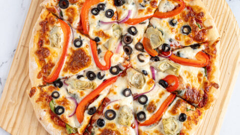 A homemade veggie pizza cut into pieces on a wooden pizza peel surrounded by pizza toppings.