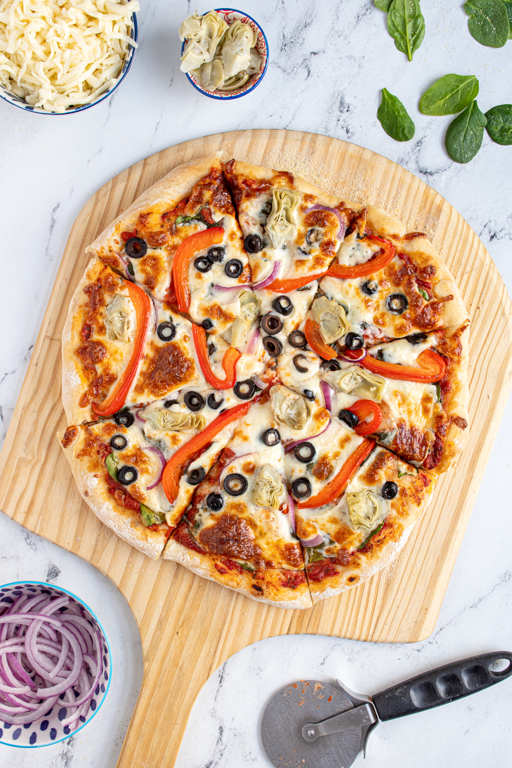 A homemade veggie pizza cut into pieces on a wooden pizza peel surrounded by pizza toppings.