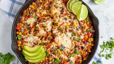 Flatlay of a black cast iron skillet on a table top with colorful corn and peppers topped with cheese and avocado.
