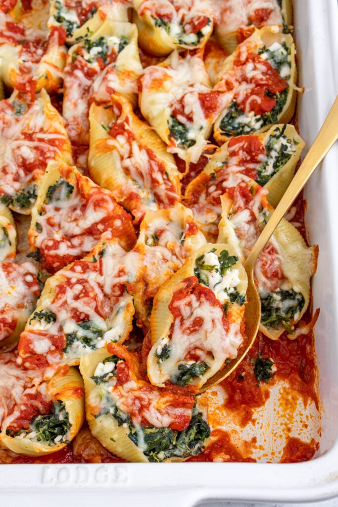 White casserole dish filled with spinach stuffed shells, a spoon taking on out of the dish.