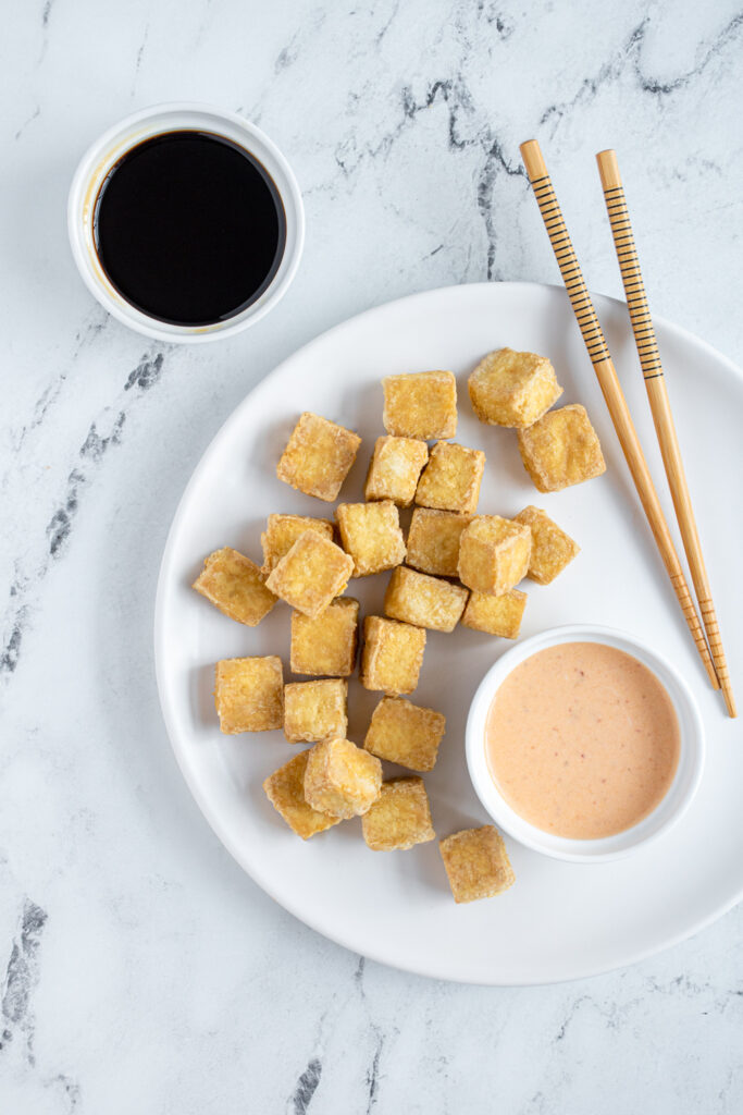 Flatlay of a white plate with dipping sauces with a pile of golden brown crispy tofu on the plate.