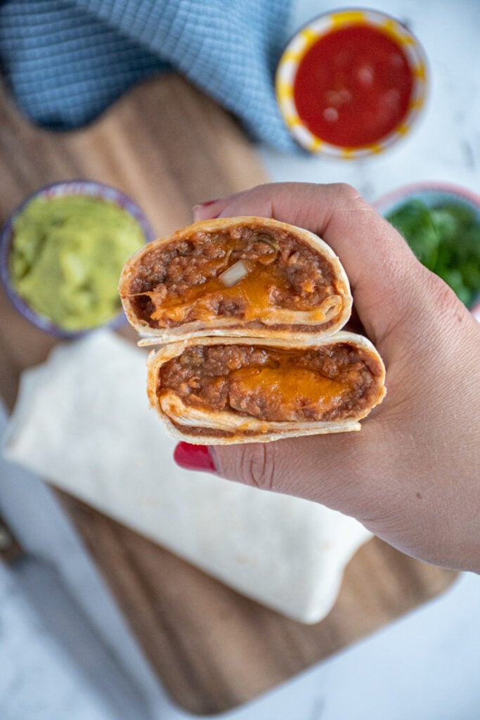 A hand holding up two halves of a bean burrito showing off the cheesy inside.
