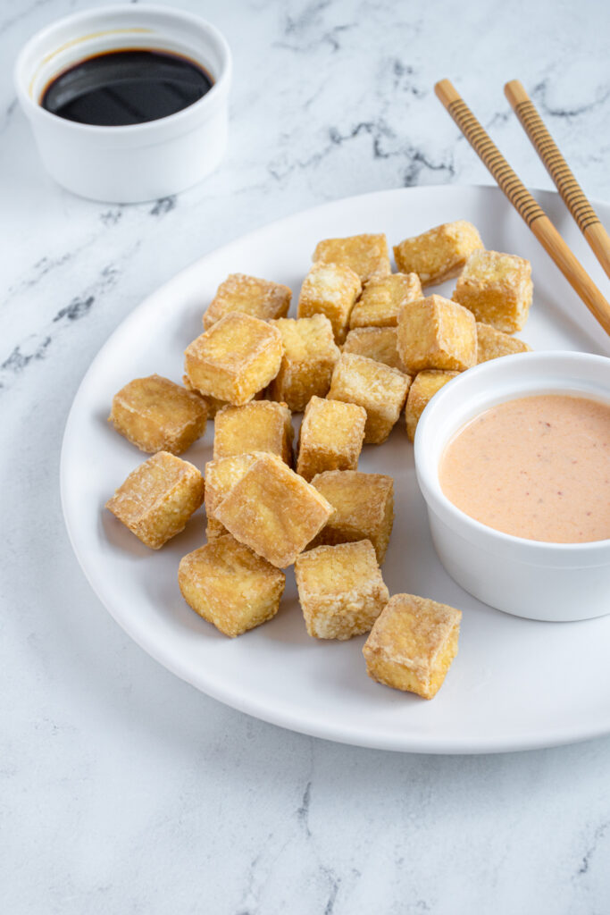 White plate with golden brown crispy air fried tofu sitting next to sauce bowls and chopsticks.
