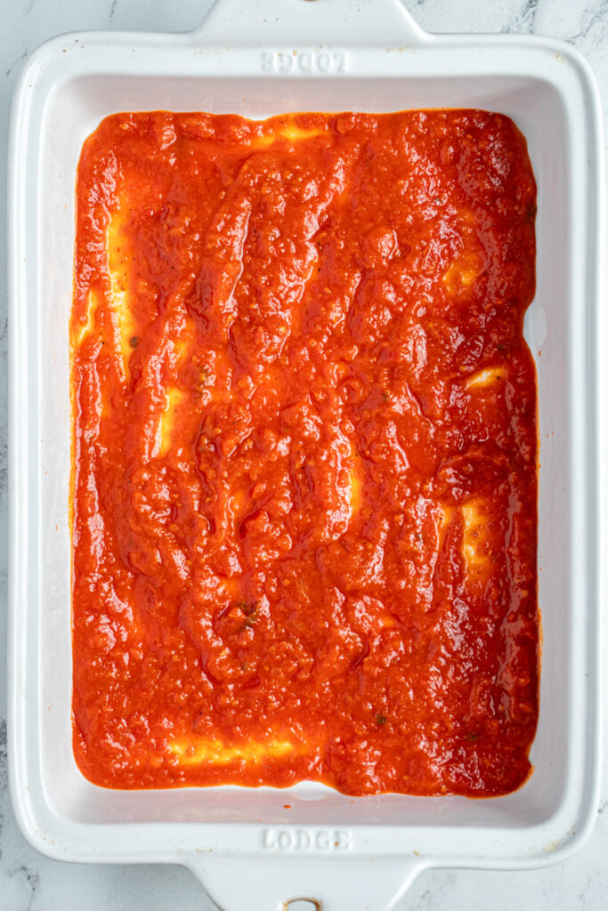 A white casserole dish with marinara sauce layered on the bottom.