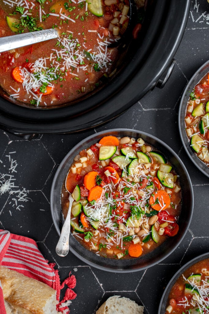 A crockpot and bowl both filled the minestrone soup garnished with parmesan cheese and parsley.