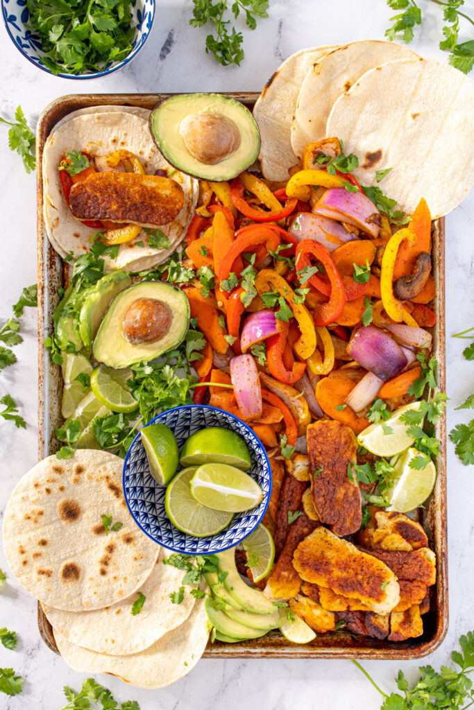 A tray filled with fajita veggies and halloumi as well as tortillas, avocados, and lime wedges all garnished with fresh cilantro.