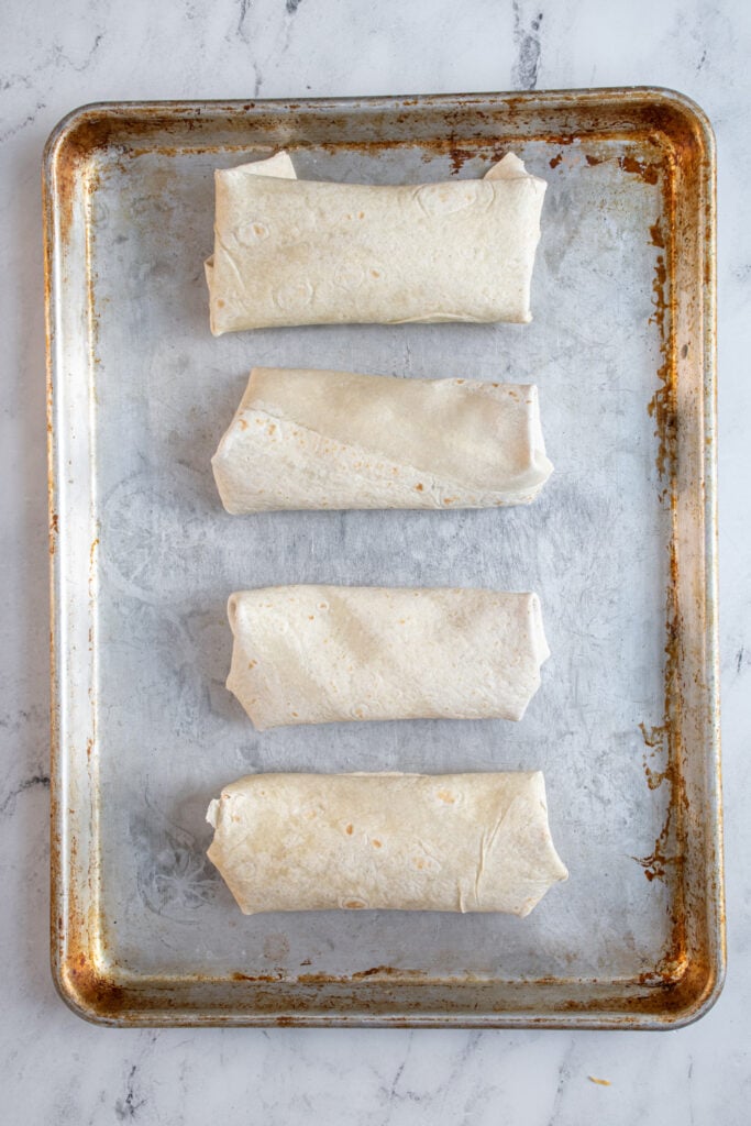 A sheet pan with four wrapped burritos ready to go into the oven.