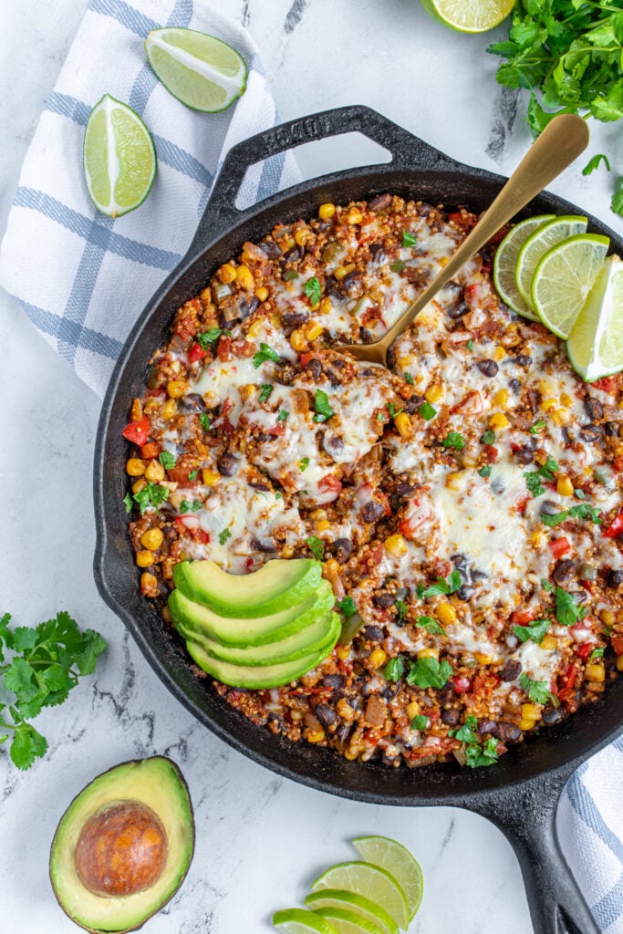 Black cast iron skillet on a tabletop with colorful corn, peppers, and beans topped with cheese and avocado slices.