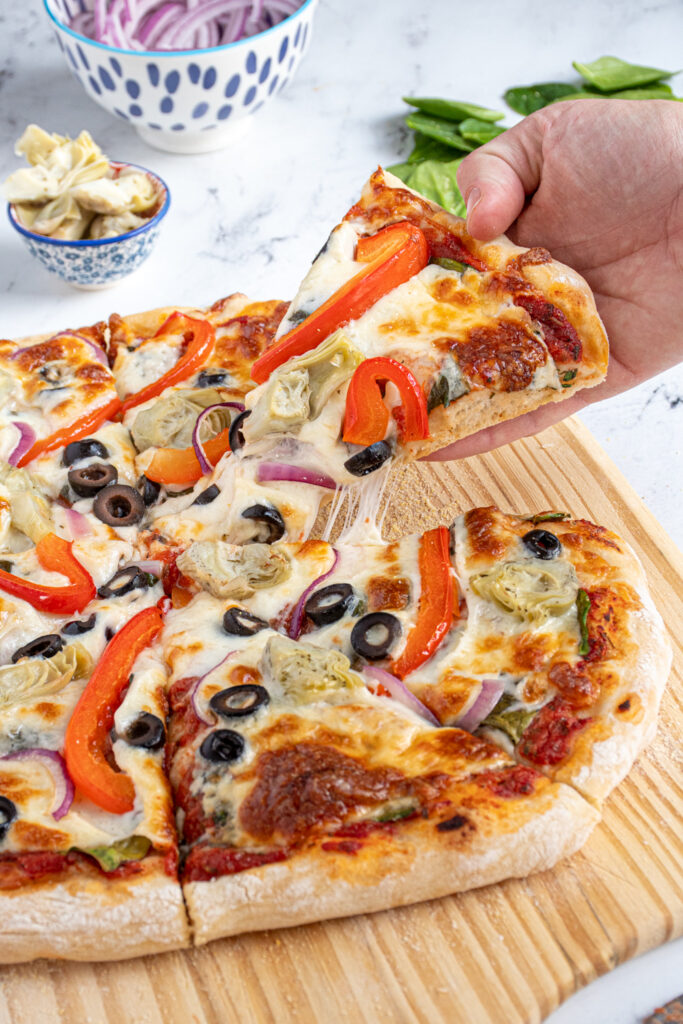 A hand pulling a slice of veggie pizza away leaving strings or gooey mozzarella.