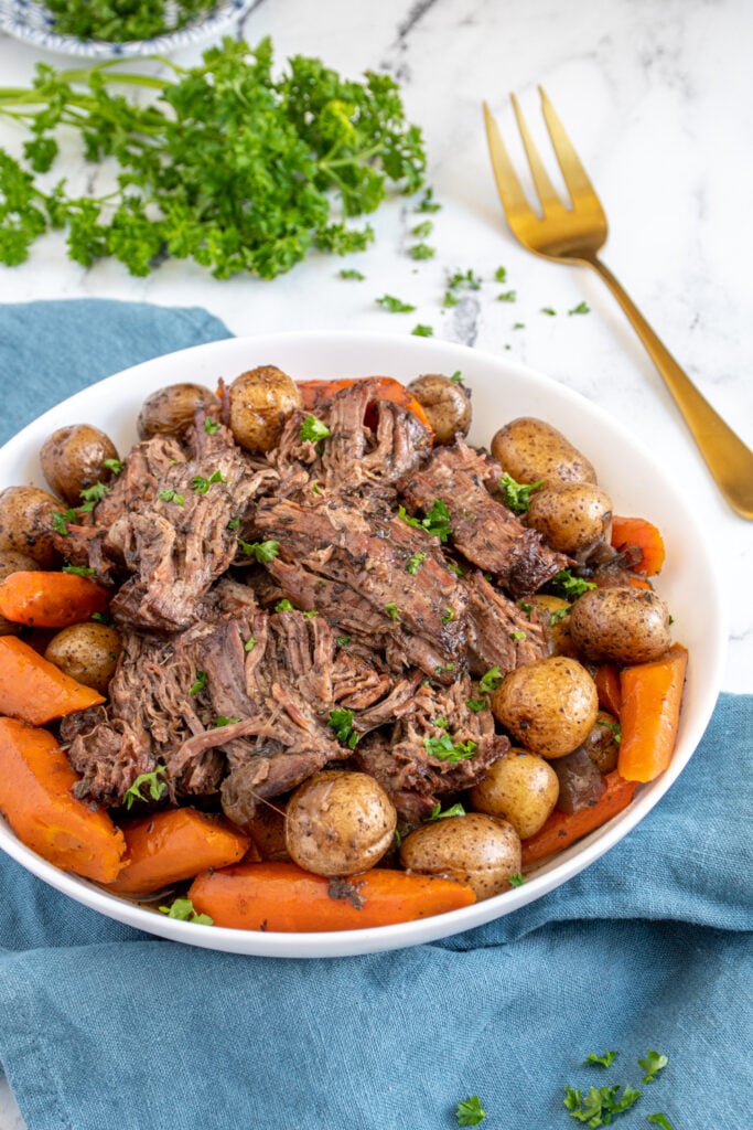 Serving bowl filled with carrots, baby potatoes, and pot roast garnished with parsley and a gold serving fork.