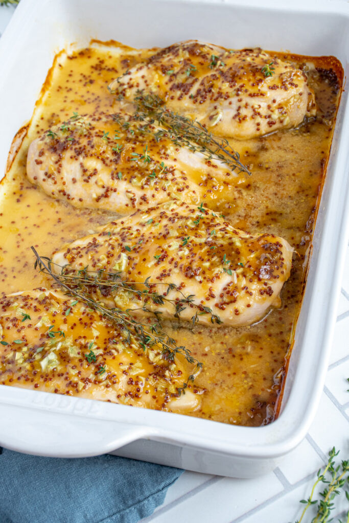 Angled white baking dish with four baked chicken breasts, covered with honey mustard sauce and garnished with fresh thyme.