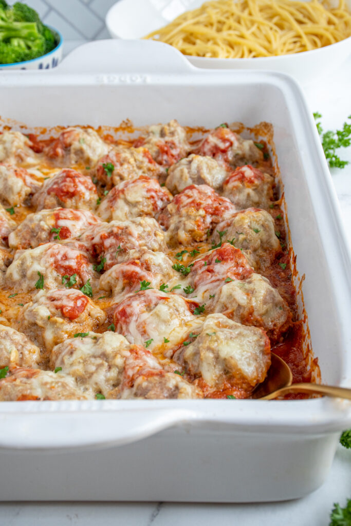 A white casserole dish at an angle filled with baked turkey meatballs in sauce and covered with mozzarella cheese and topped with parsley.