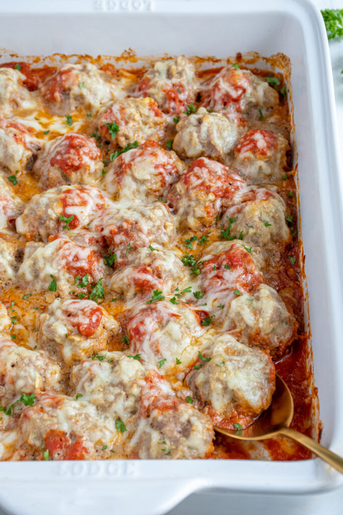 Close up of baked turkey meatballs in a white casserole dish with marinara sauce and covered in cheese and garnished with parsley.