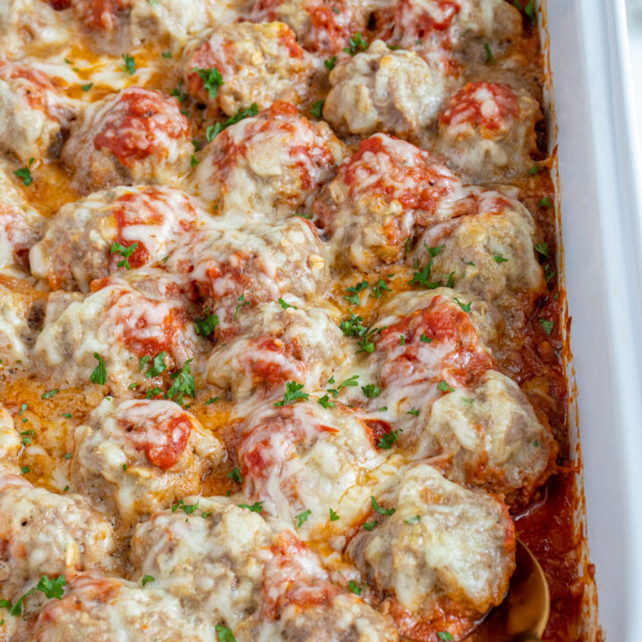Close up of baked turkey meatballs in a white casserole dish with marinara sauce and covered in cheese and garnished with parsley.