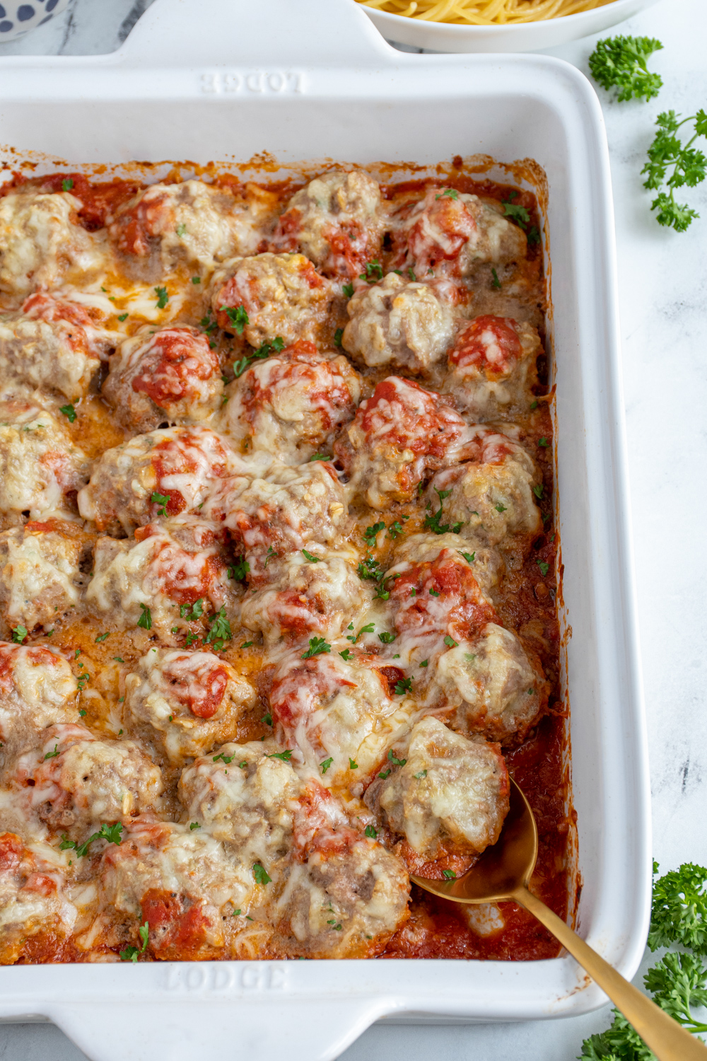 White casserole dish with baked turkey meatballs in sauce covered with cheese and fresh parsley.