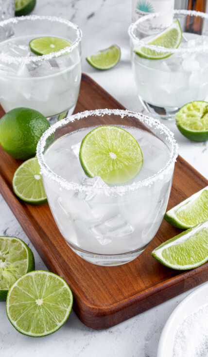 A classic margarita on a wooden platter surrounded by lime halves with a salted rim and a lime wheel as garnish.