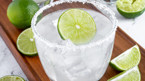 A classic margarita on a wooden platter surrounded by lime halves with a salted rim and a lime wheel as garnish.