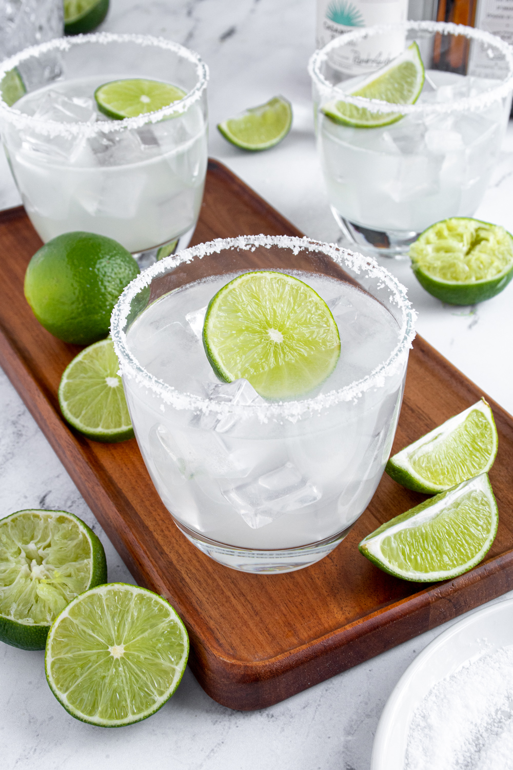 A classic margarita on a wooden platter surrounded by lime halves with a salted rim and a lime wheel as garnish.