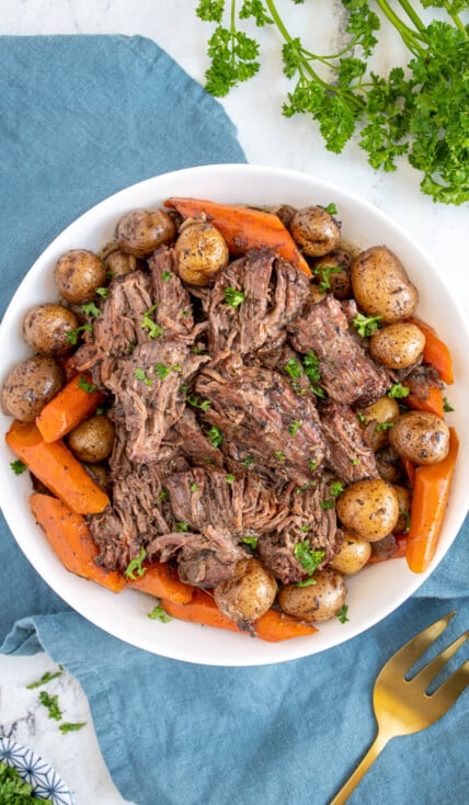 Large white serving bowl filled with carrots, baby potatoes, and juicy pot roast chunks garnished with little specks of parsley.