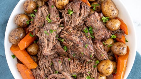 Large white serving bowl filled with carrots, baby potatoes, and juicy pot roast chunks garnished with little specks of parsley.