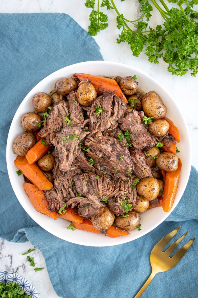 Large white serving bowl filled with carrots, baby potatoes, and juicy pot roast chunks garnished with little specks of parsley.