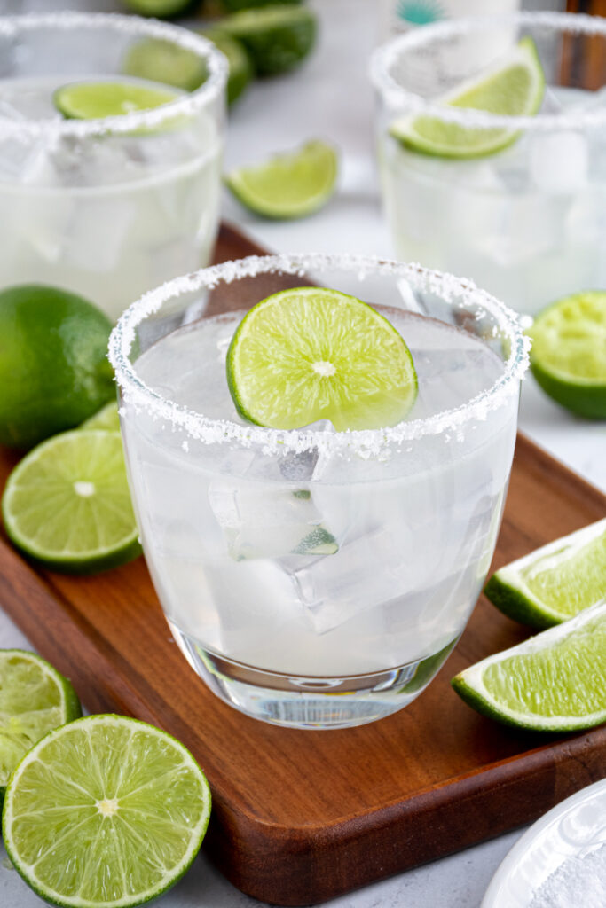 A glass with a salted rim filled with a margarita and garnished with a lime wheel on a wood platter.