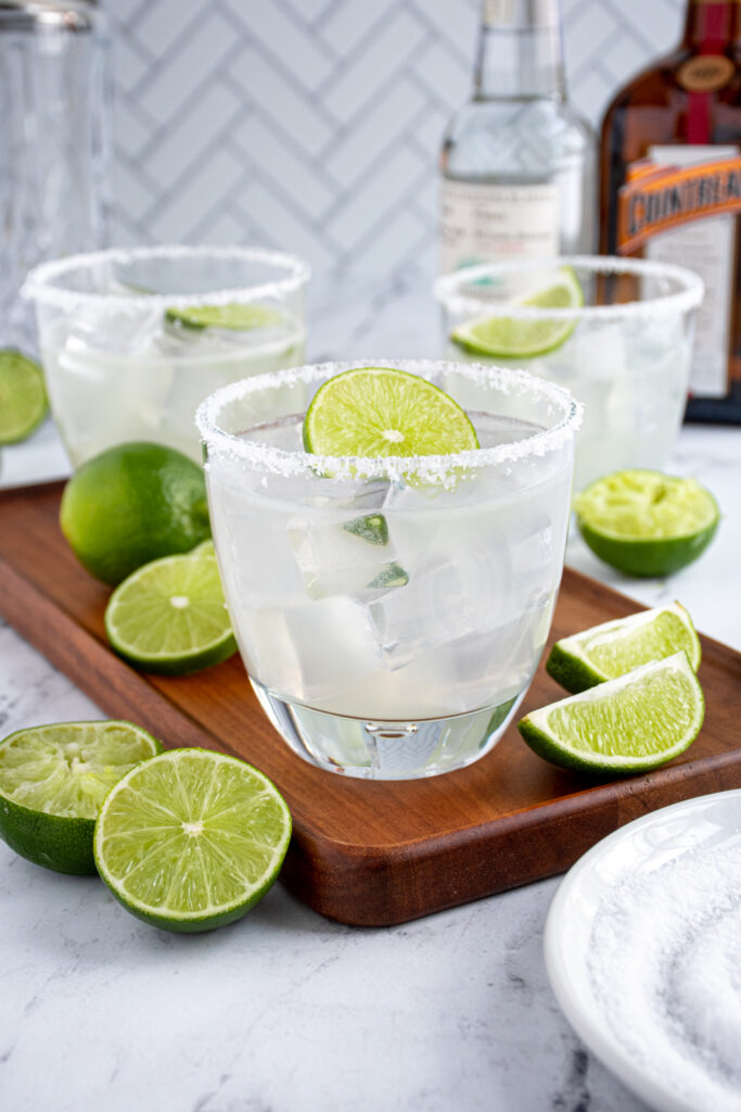 Three classic margaritas with salted rims and lime garnishes on a table top with bottles and lime halves.