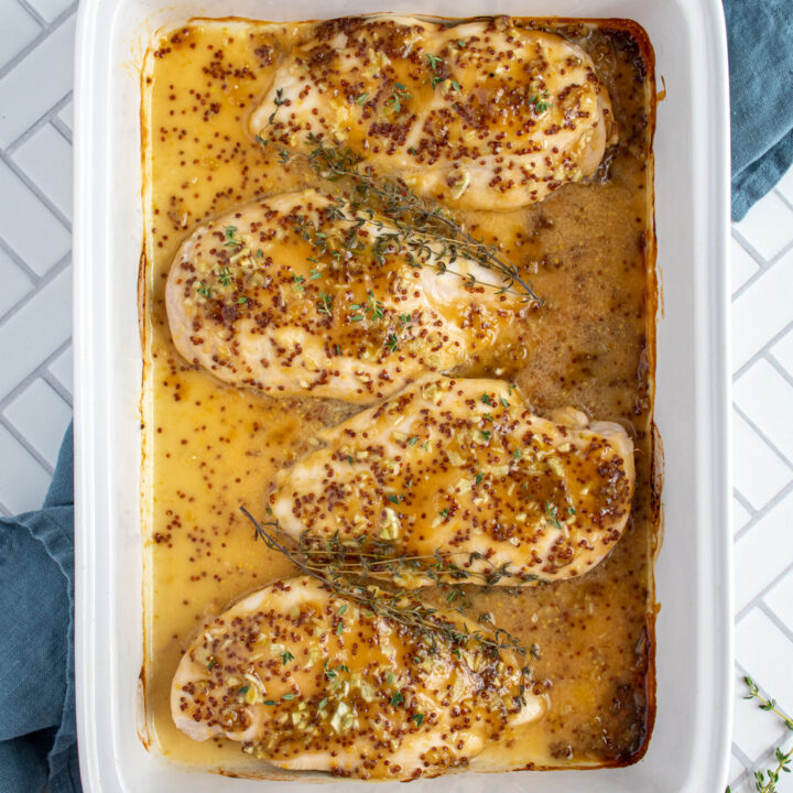 Overhead view of a white baking dish filled with four chicken breasts with a homemade honey mustard sauce overtop and garnished with fresh thyme.