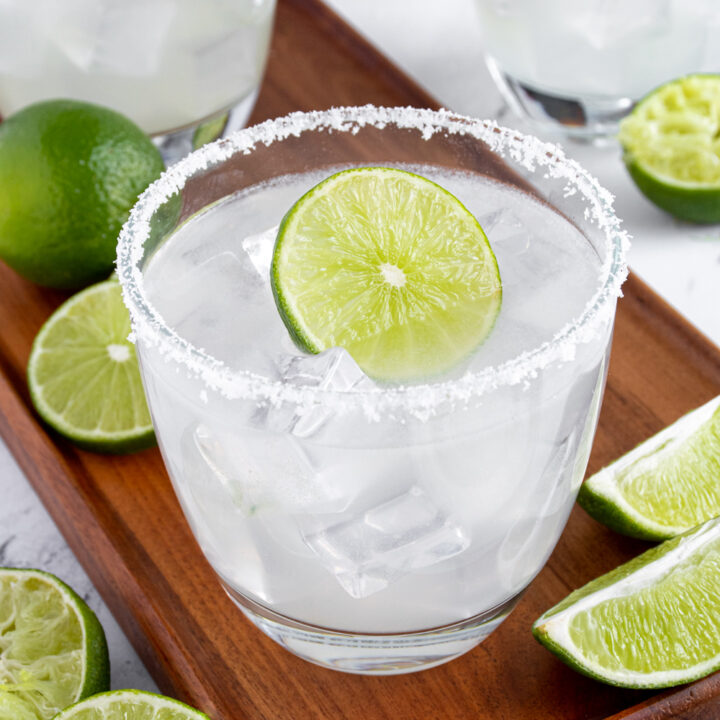 A glass with a salted rim filled with ice and margarita garnished with a lime wheel, sitting on a platter with other drinks and limes in the background.