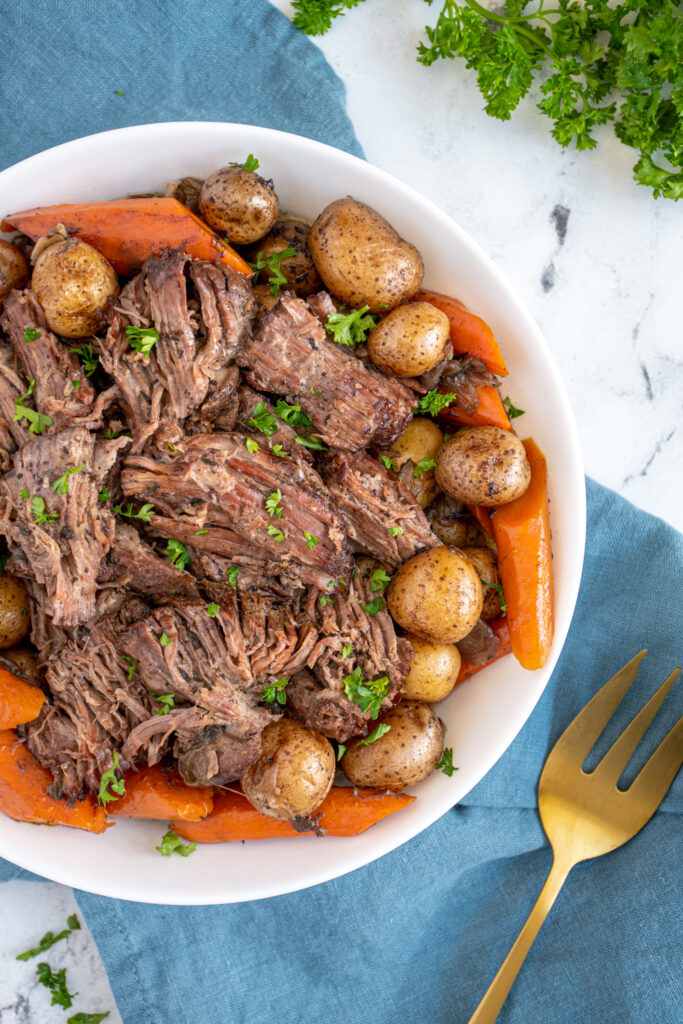 A white serving bowl filled with crockpot pot roast with red wine, baby potatoes, and carrots all garnished with fresh parsley.