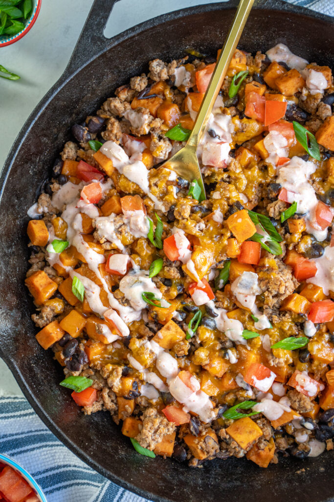 Close up of a cast iron skillet with ground turkey, sweet potatoes, and black beans, topped with diced tomatoes, green onions, and a sauce drizzle.