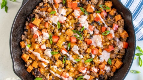 Cast iron skillet holding ground turkey and sweet potato skillet with black bean garnished with diced tomatoes, sliced green onions, and a drizzle of sauce.