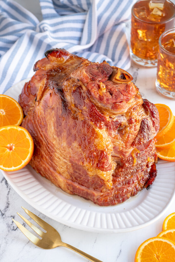 A ham with orange bourbon glaze on a white platter garnished with orange slices.