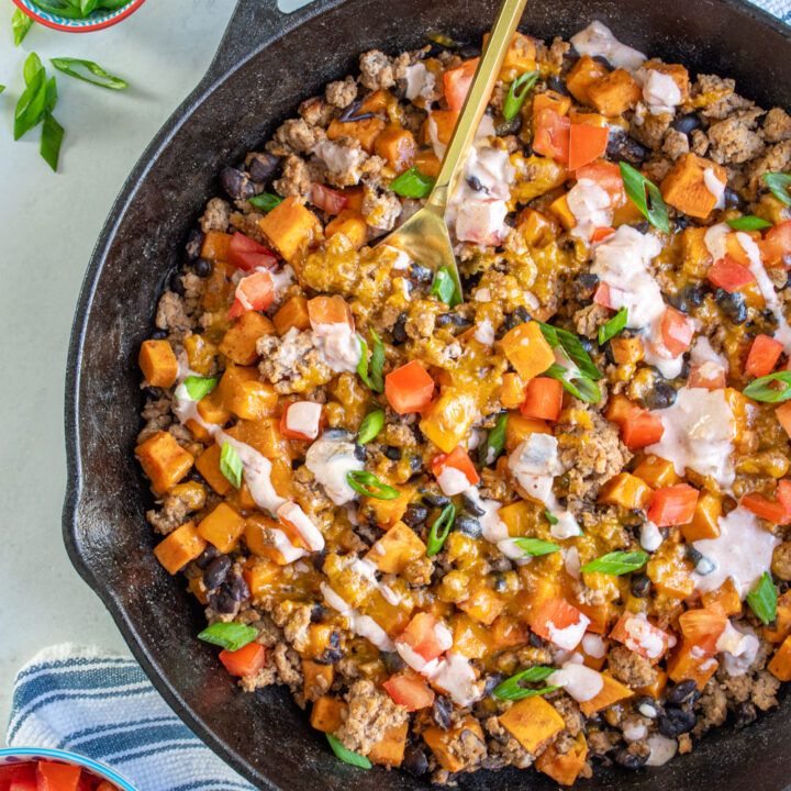 Cast iron pan with ground turkey sweet potato skillet with black beans and garnished with diced tomatoes, green onions, and a drizzle of a sauce.