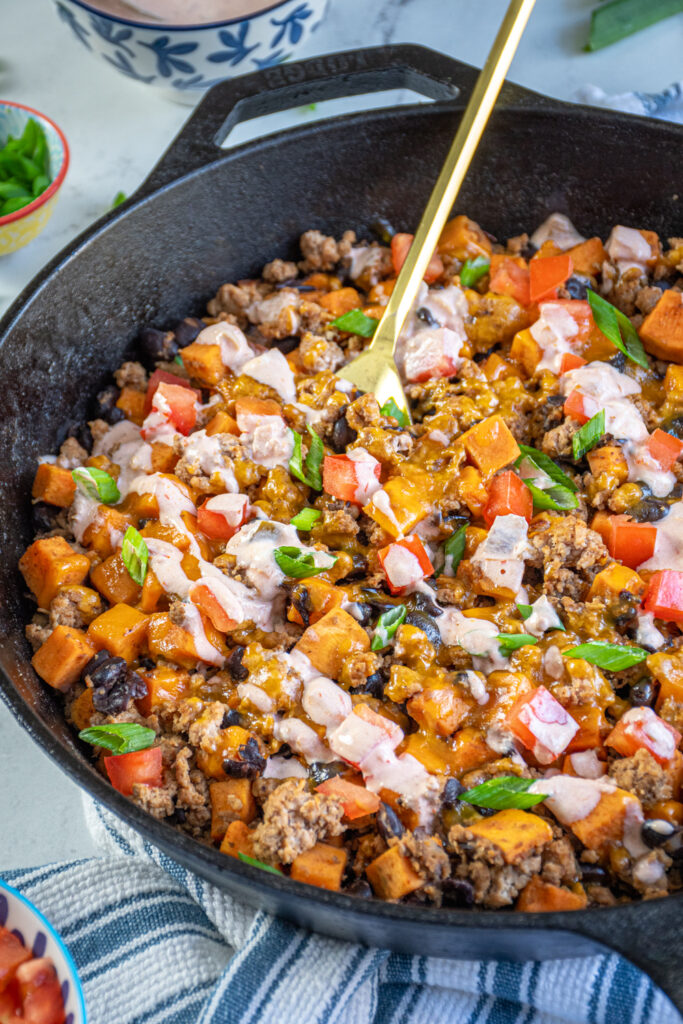 A gold serving spoon in a turkey and sweet potato skillet with black beans and garnished with diced tomatoes and a drizzle of sauce.