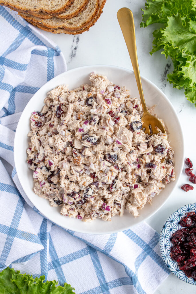 Bowl of tuna salad with cranberries with a gold spoon surrounded by lettuce and bread.