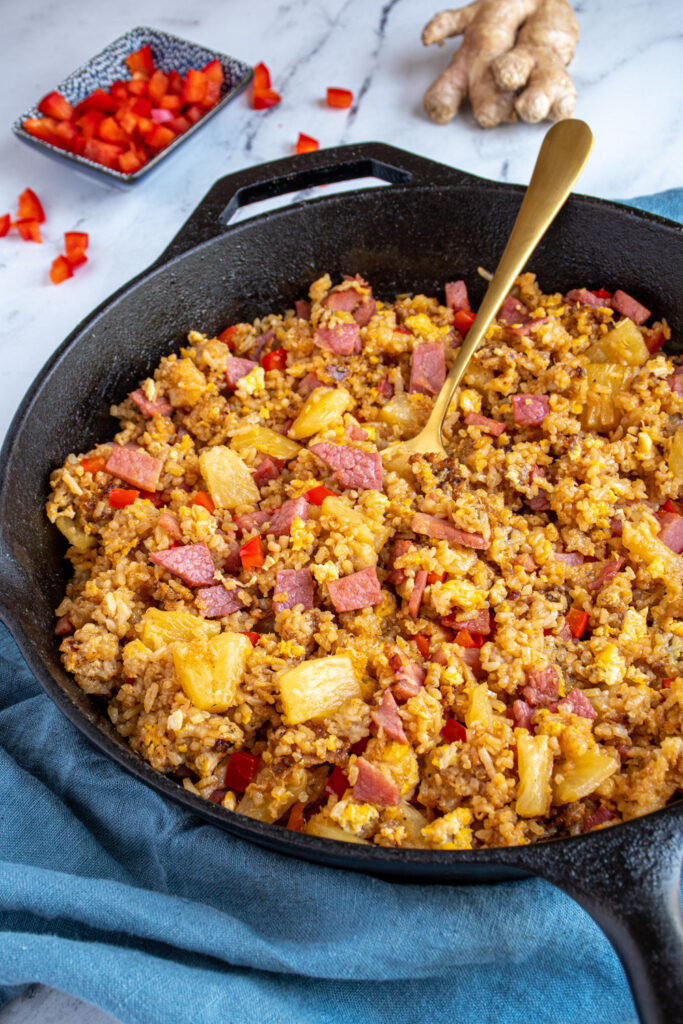 A skillet with a serving spoon in ham fried rice with pineapple and red bell pepper.