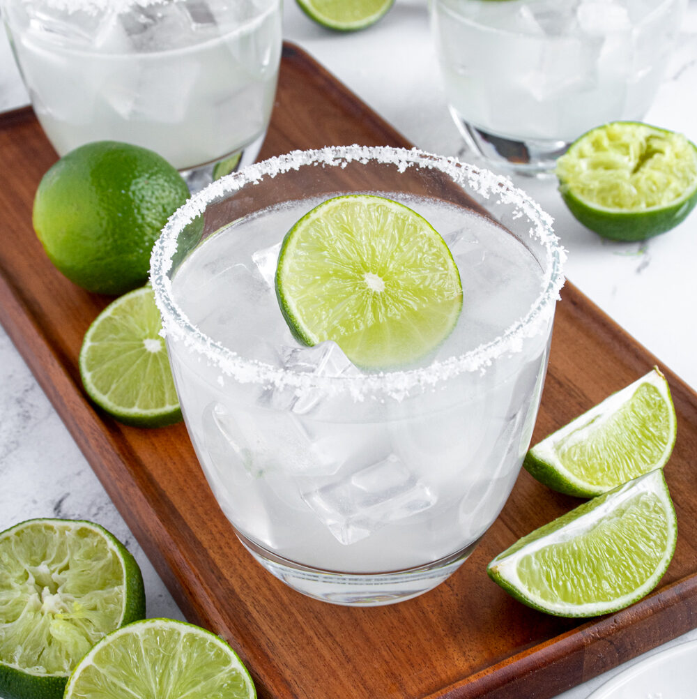 A classic margarita on a wooden platter surrounded by lime halves with a salted rim and a lime wheel as garnish.