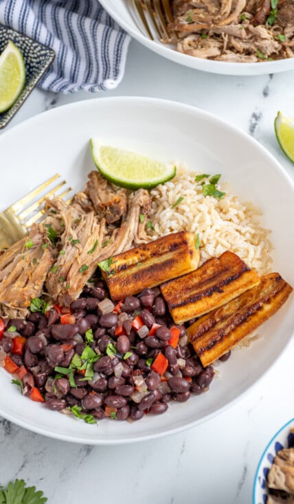 A cuban bowl filled with rice, mojo pork, cuban black beans, and fried bananas garnished with cilantro and lime wedges.