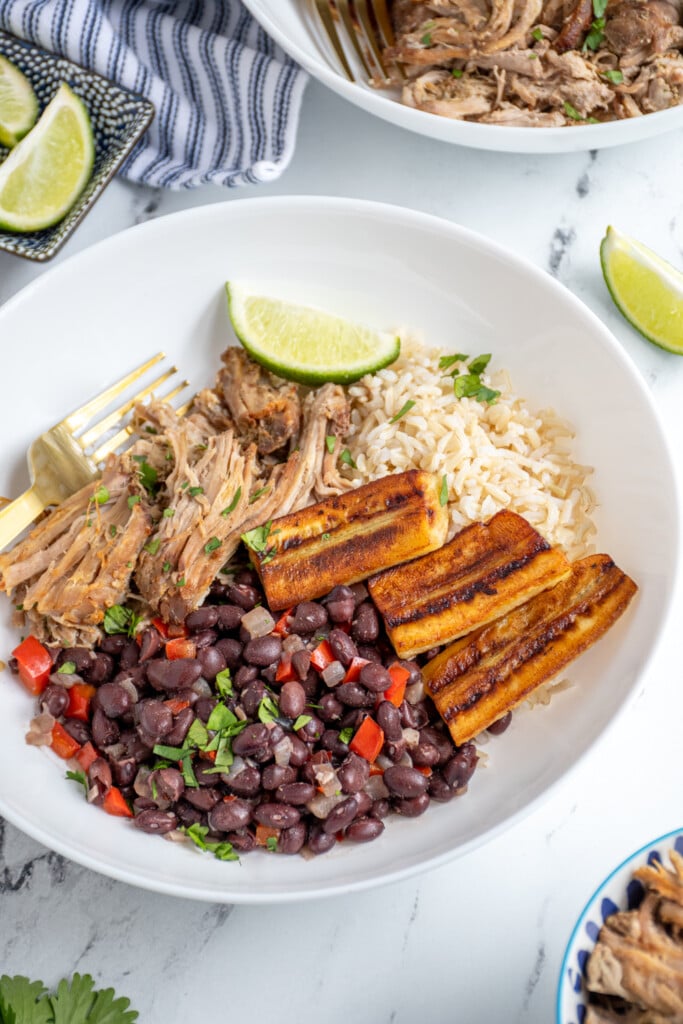 A cuban bowl filled with rice, mojo pork, cuban black beans, and fried bananas garnished with cilantro and lime wedges.