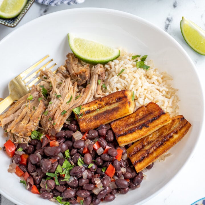 A cuban bowl filled with rice, mojo pork, cuban black beans, and fried bananas garnished with cilantro and lime wedges.