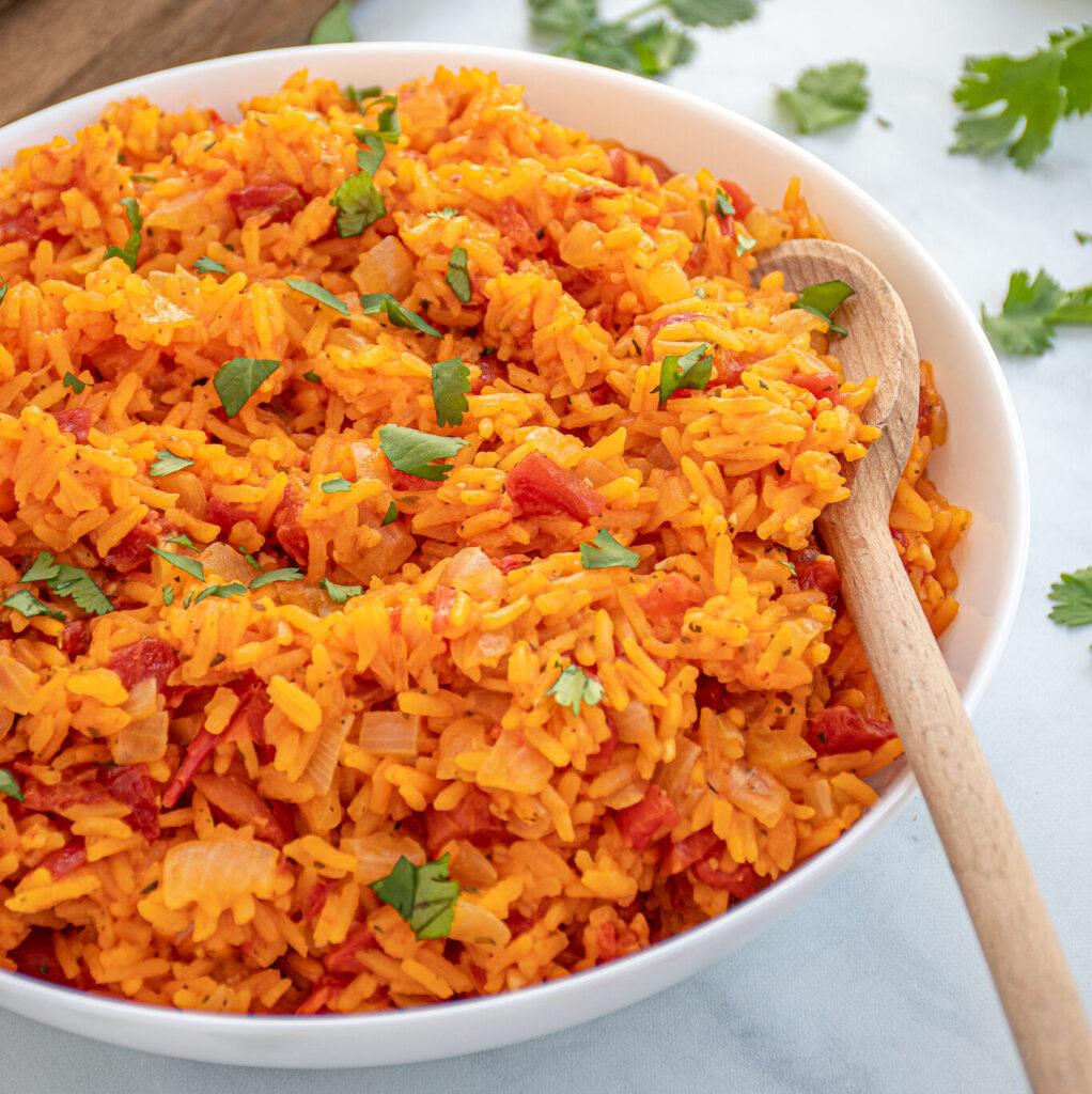 Bright orange-red colored Mexican Rice in a large white bowl with a wooden spoon garnished with chopped cilantro.