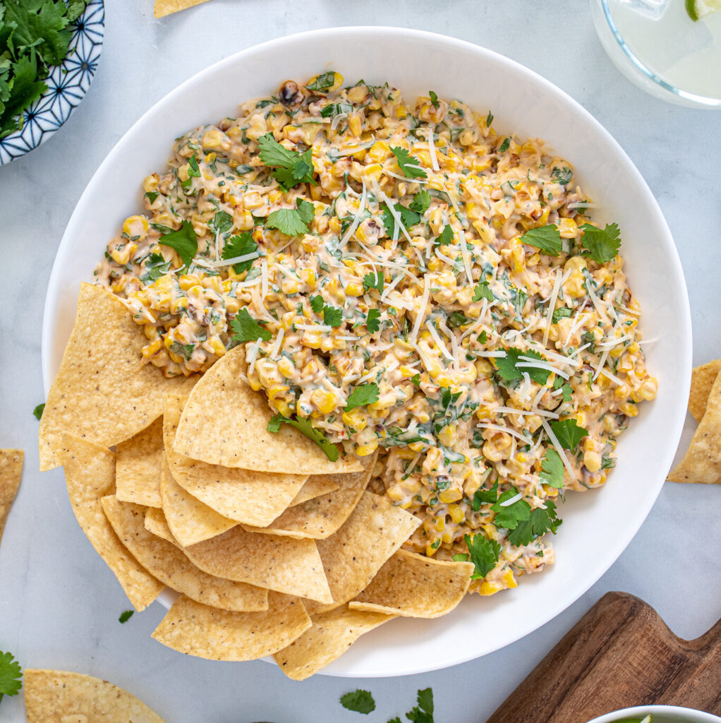 Large what bowl filled with Mexican Street Corn Dip and tortilla chips surrounded by more chips, cilantro, and limes.