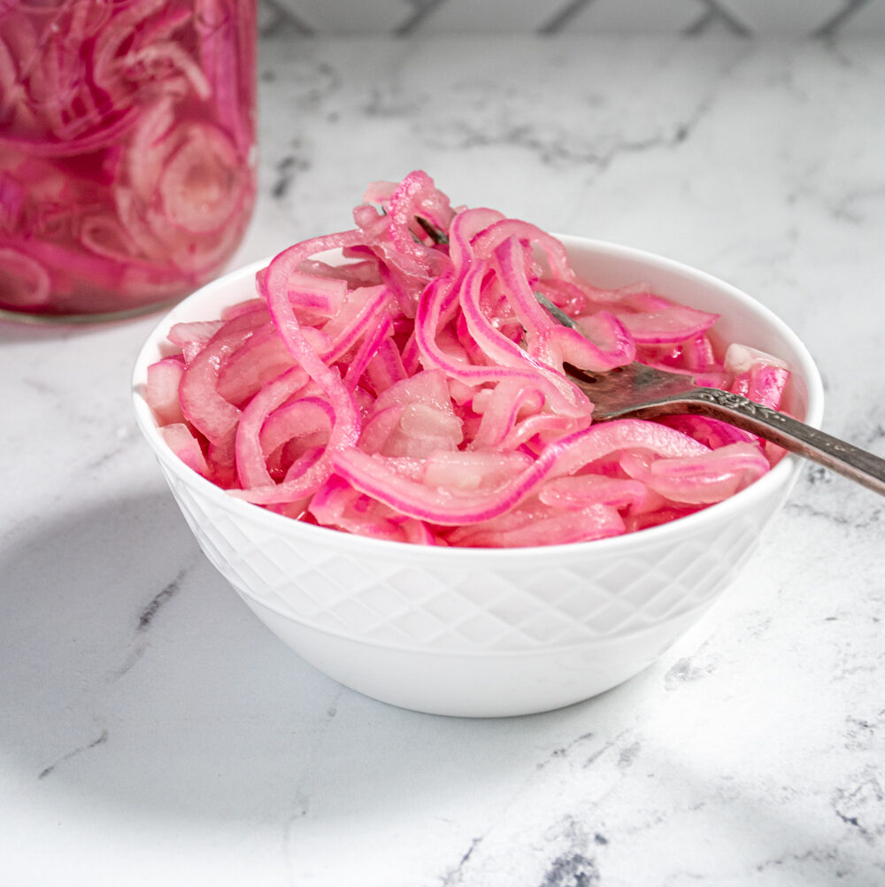 A white bowl with quick pickled red onions and a fork on a countertop next to a jar full of quick pickled onions.