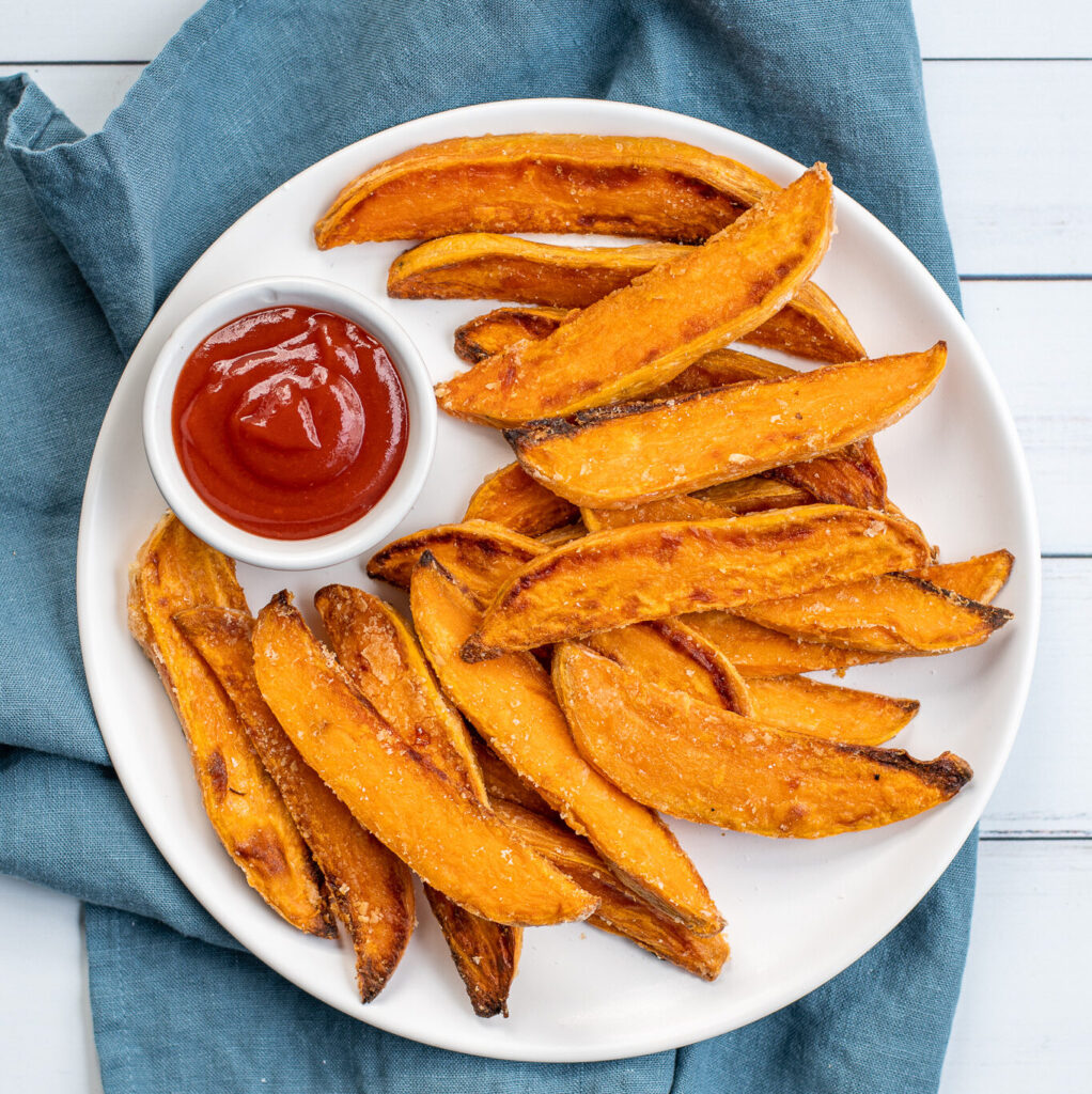 Overhead shot of a white plate sitting on blue towel the plate in piled high with crispy sweet potato wedges and a little cup of ketchup.